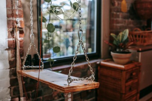 Wooden swing with chains near window and potted plant at home with brick wall