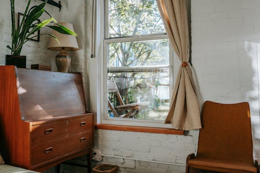 A sunlit room with vintage furniture, featuring a wooden cabinet, chair, and window view.