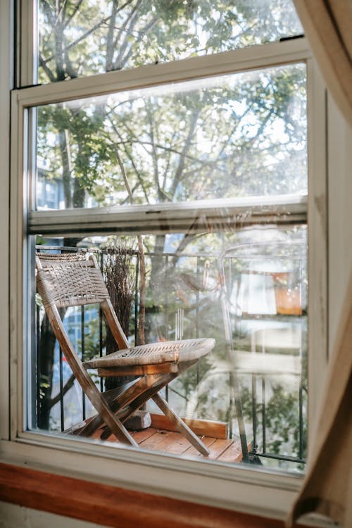 Through apartment window glass view of rustic wooden chair placed on small balcony on sunny day