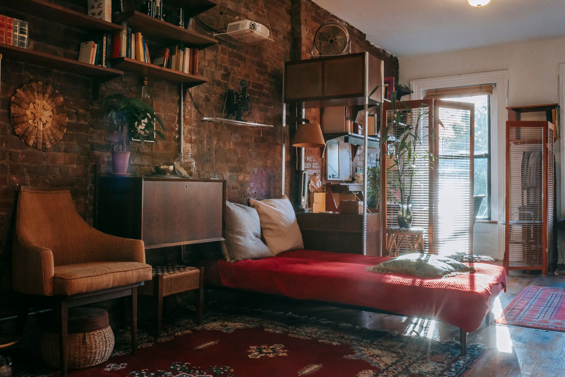 Warm book-lined bedroom with dark wood furniture, red bedding, weathered brick, patterned rugs and a reading chair