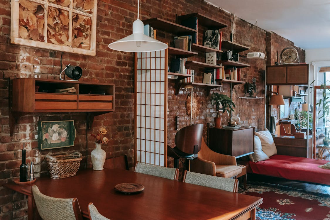 Free Interior of cozy vintage styled dining room with classic wooden table and chairs placed near weathered brick wall decorated with paintings and bookshelves Stock Photo