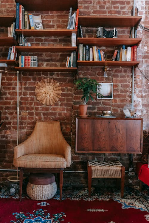 Stack of books placed on shelves attached to brick wall in cozy room with classic wooden furniture and comfortable armchair