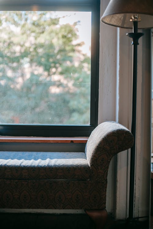 Small soft coach placed near ornamental lamp and window in cozy apartment in daytime