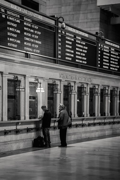 Black and white of faceless people standing near ticket window in railway station with classic interior