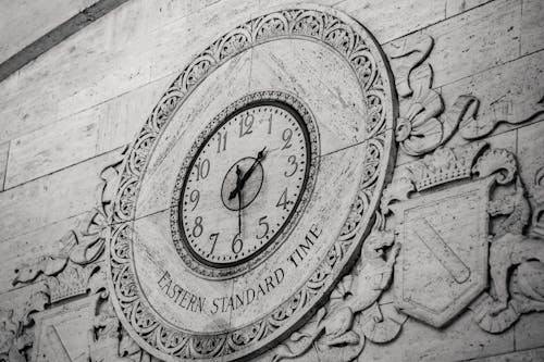 From below black and white of stone wall with ornamental details around clock showing Eastern Standard Time