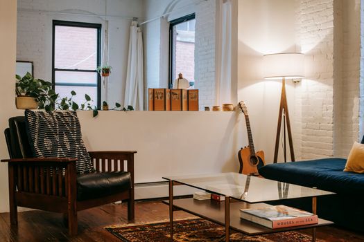 Modern room with sofa and guitar placed near glass table and soft armchair in living room with potted plant and windows