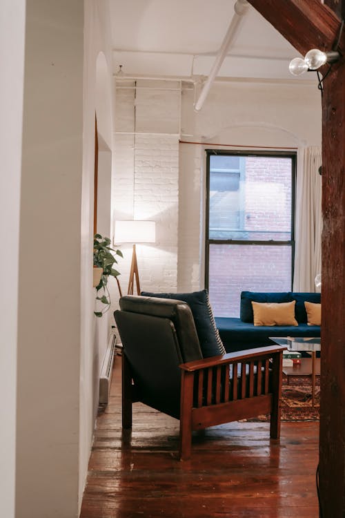 View of cozy living room with armchair and sofa near window through doorway decorated with lamps in modern apartment with white walls