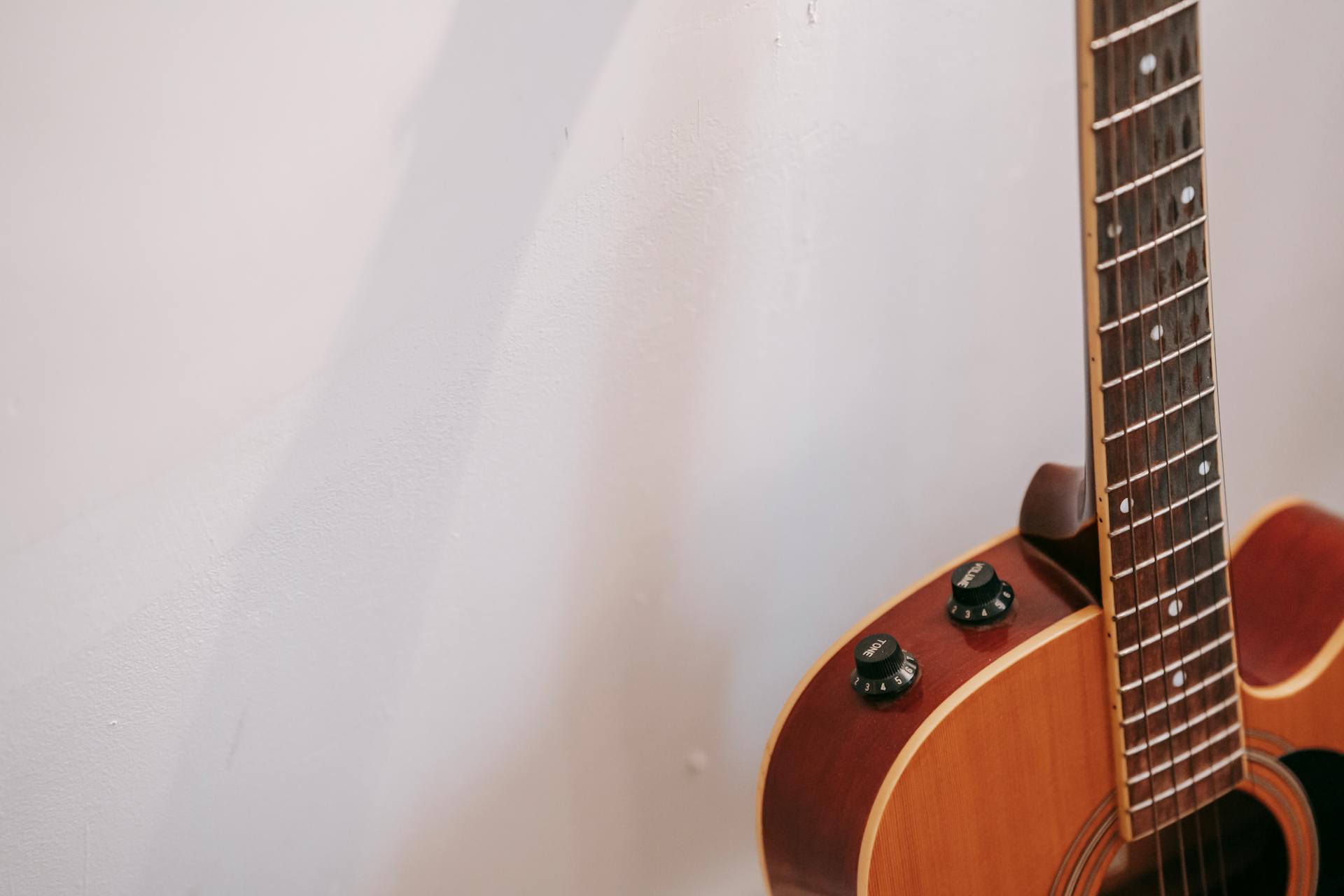 Detail of guitar neck with volume and tone knobs on body of brown musical instrument placed near white wall with shadow