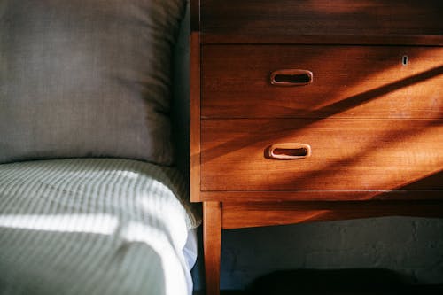 Free Interior of contemporary cozy bedroom including brown wooden night stand near bed covered with blanket and soft pillow in sun rays Stock Photo