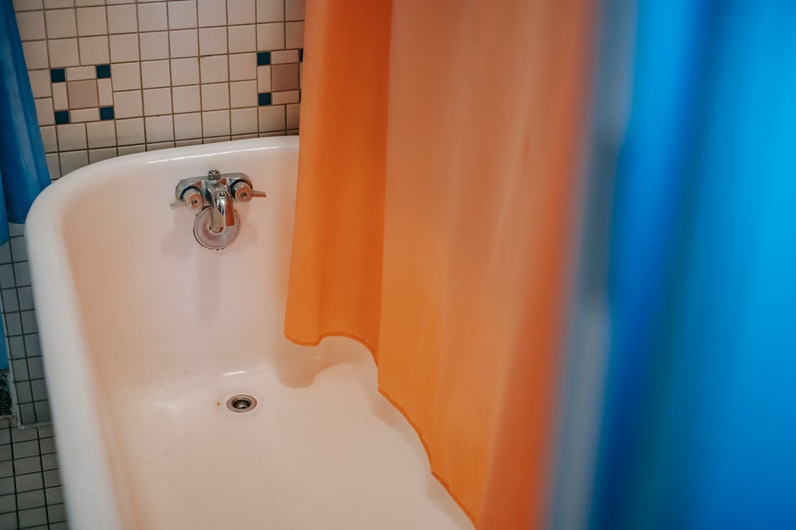 From above of clear white bath with steel faucet with hanging blue and orange curtain located in bathroom with tiled walls