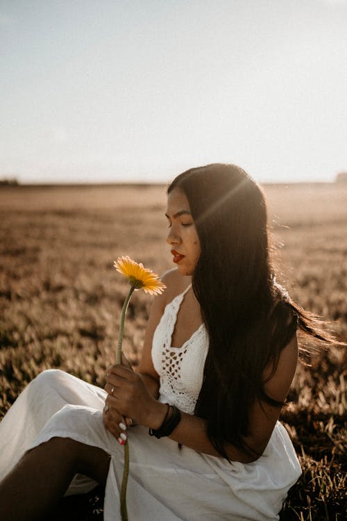 Young tender ethnic female in white sundress with blossoming flower resting on meadow in countryside in sunshine