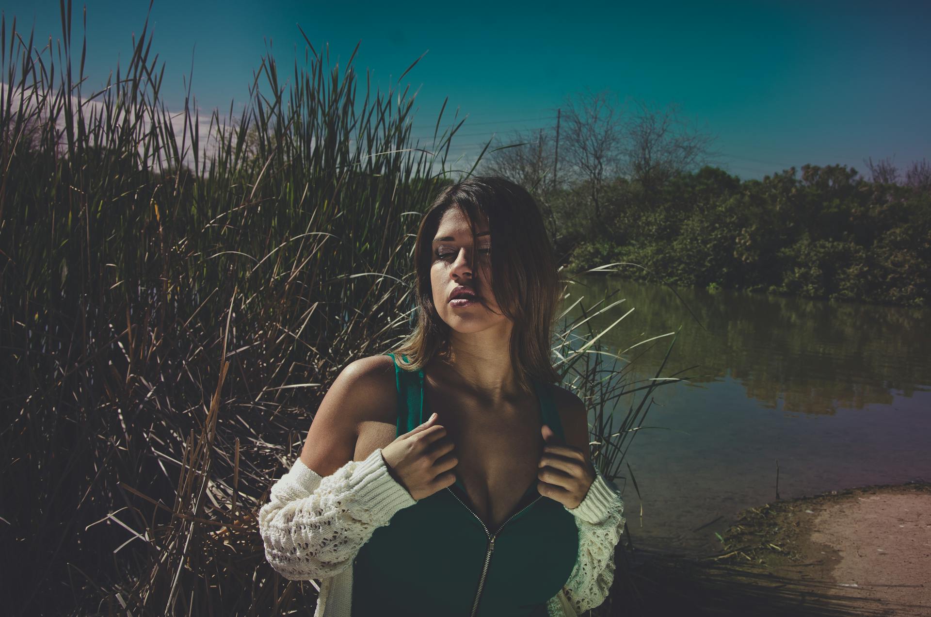 Sensual young female in swimwear and cardigan standing on shore near calm lake and green bushes in summer sunny day