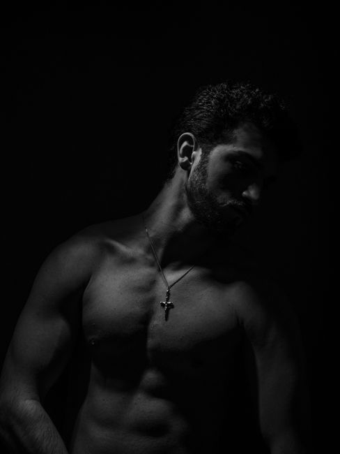 Artistic black and white portrait of a shirtless, muscular man with a cross necklace in a studio shoot.