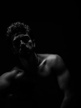 Dramatic black and white portrait of a shirtless man in a dark studio setting, highlighting muscular definition.