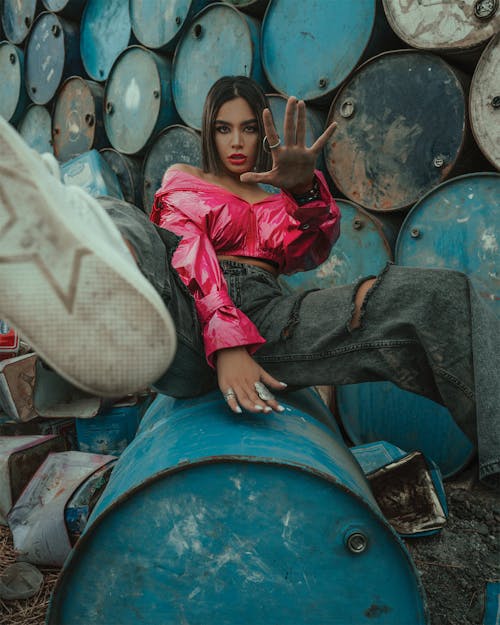 Ethnic cool female with dark hair in stylish outfit outstretching hand at camera sitting on blue barrel in dim light