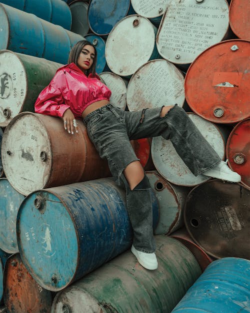 Full body of calm ethnic female with dark hair in trendy bright clothes lying on old barrels in dim daylight