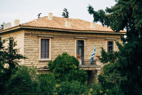 Free Historic stone villa with a Greek flag surrounded by greenery, showcasing classic architecture and lush landscape. Stock Photo