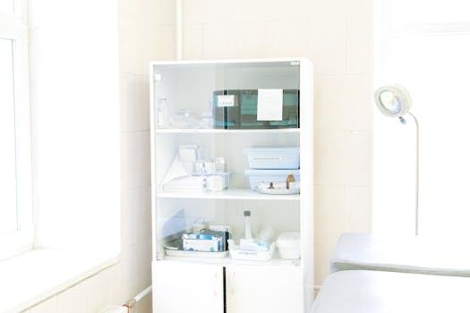 Modern white cabinet with cupboard with various medical instruments and shelves with medical materials near empty white bed near window and lamp in hospital