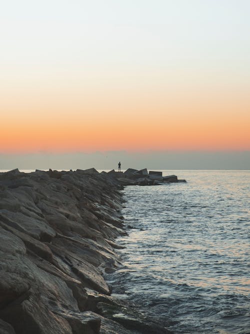 A Silhouette of a Person Standing on Rock Formation Near Body of Water during Sunset