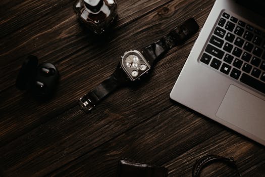 A sleek still life photo featuring a laptop, wristwatch, earphones, and a bottle on a wooden surface.