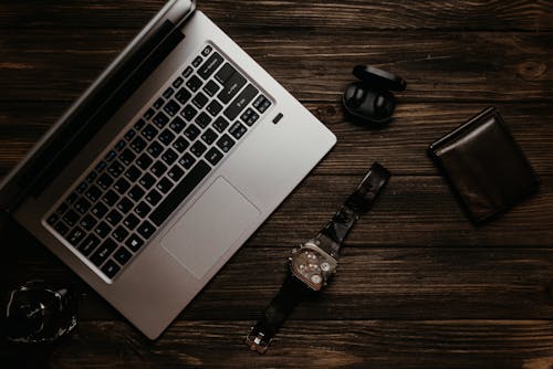 Silver Laptop Beside a Chromed Analog Watch on Brown Wooden Table