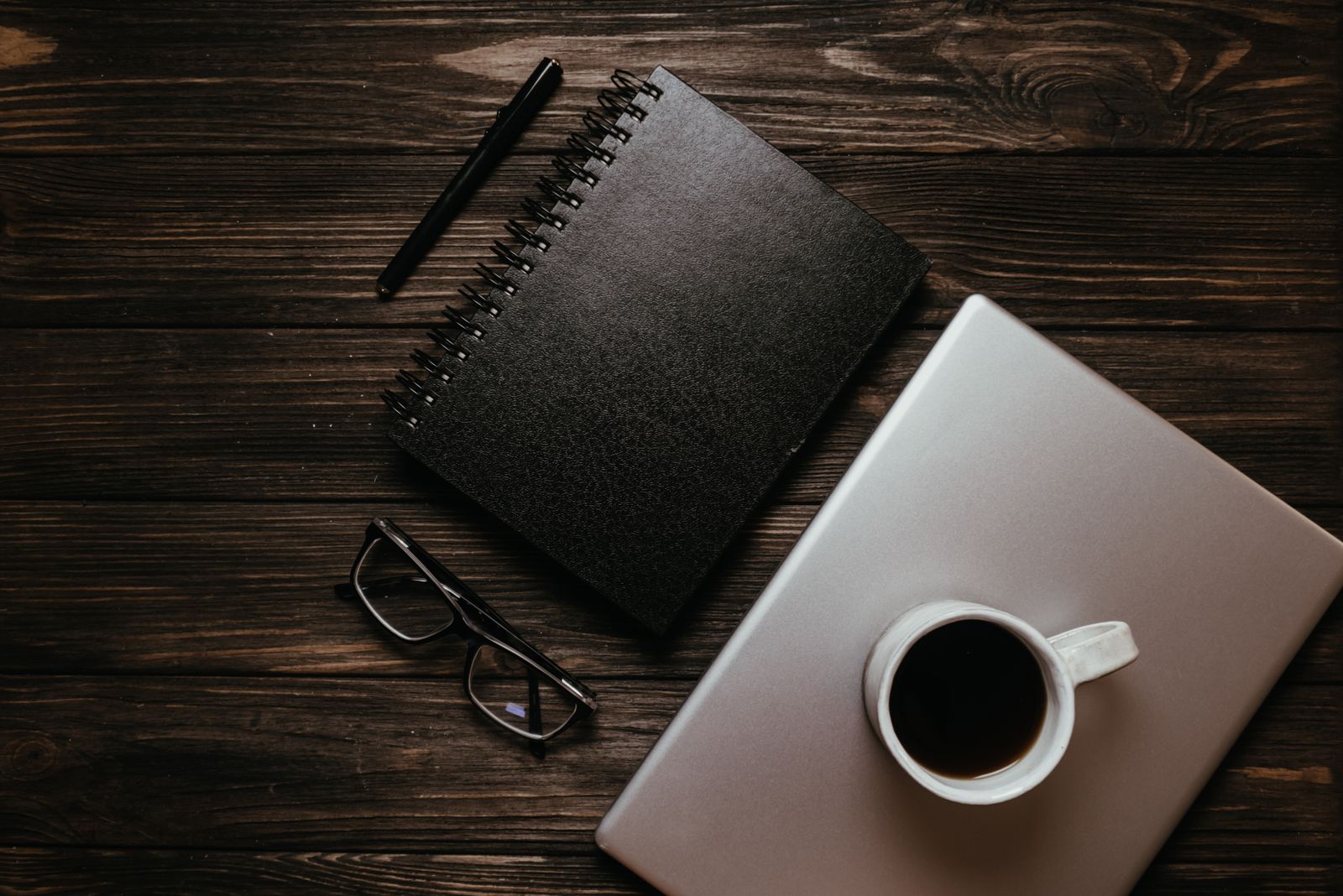 Dark wooden desk with a cup of coffee, notebook and laptop — photo by Polina on Pexels