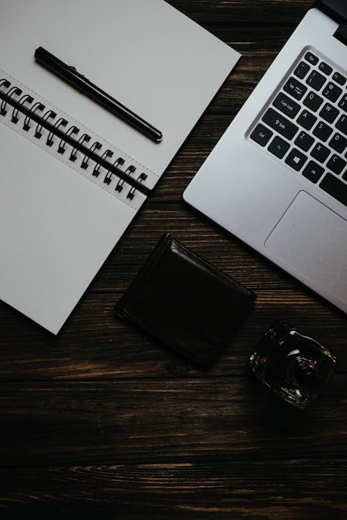 White Notebook with a Black Pen Beside a Laptop on Brown Wooden Table