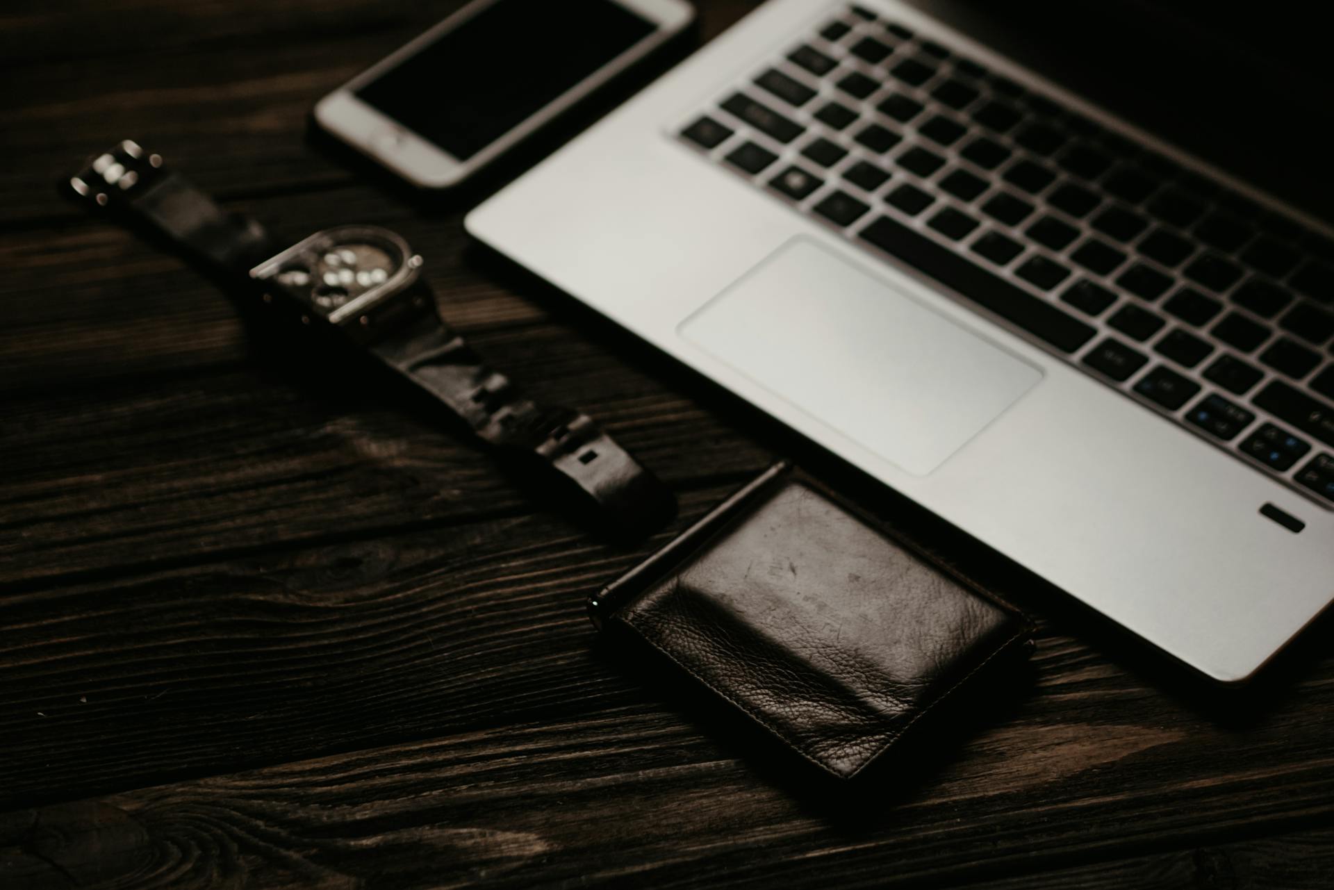 A flat lay of a laptop, wristwatch, smartphone, and wallet on a wooden table.