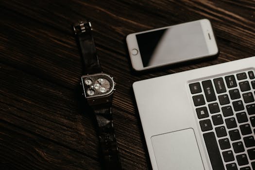 Close-up of a laptop, smartphone, and wristwatch on a wooden table, capturing a sleek workspace setup.