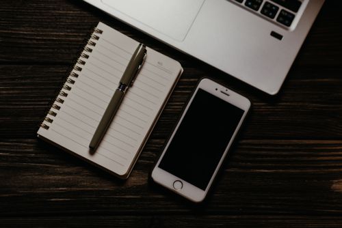 Free Close-up of a modern workspace featuring a notebook, pen, smartphone, and laptop on a wooden table. Stock Photo