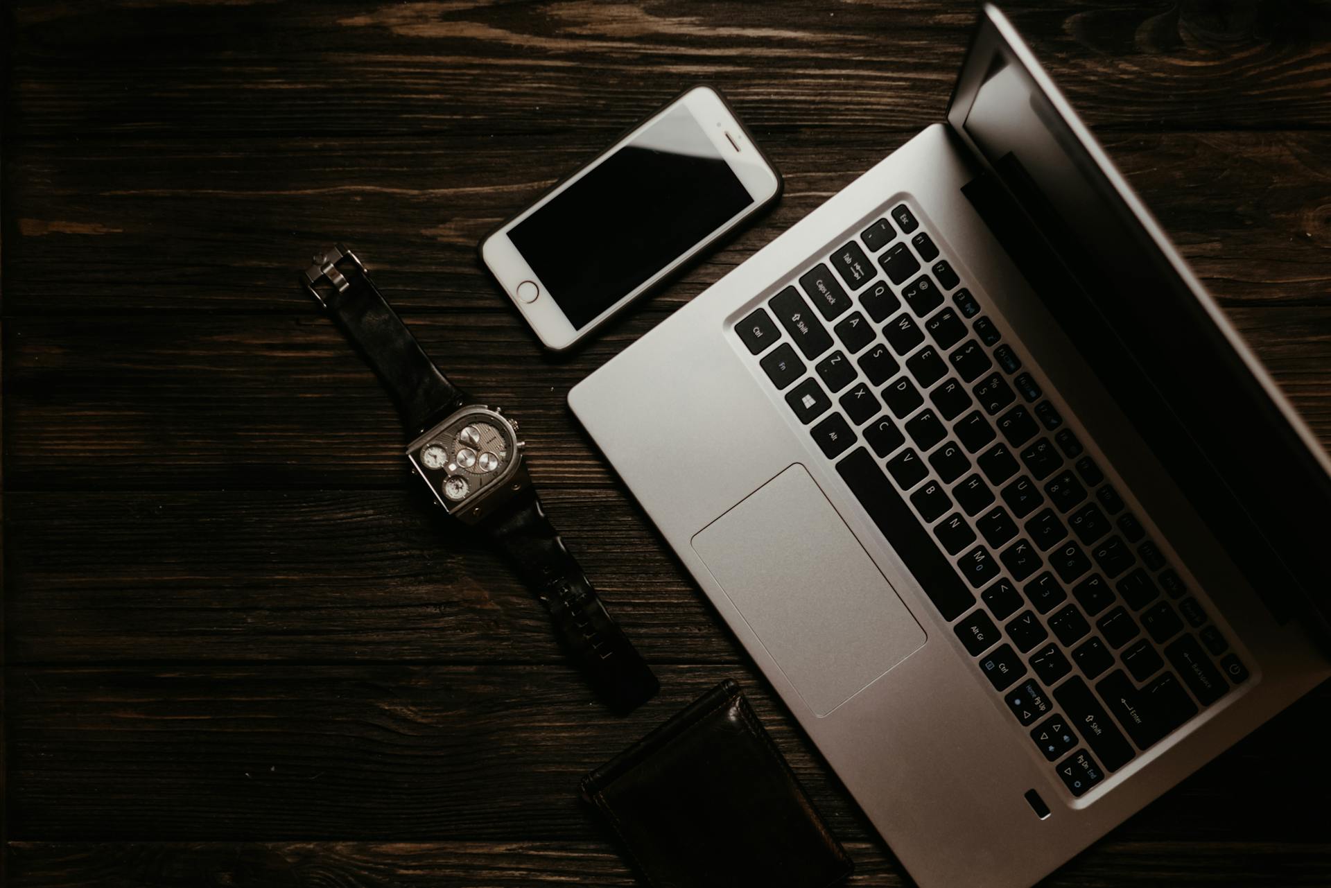 Top view of laptop, smartphone, wristwatch, and wallet on a dark wooden surface.