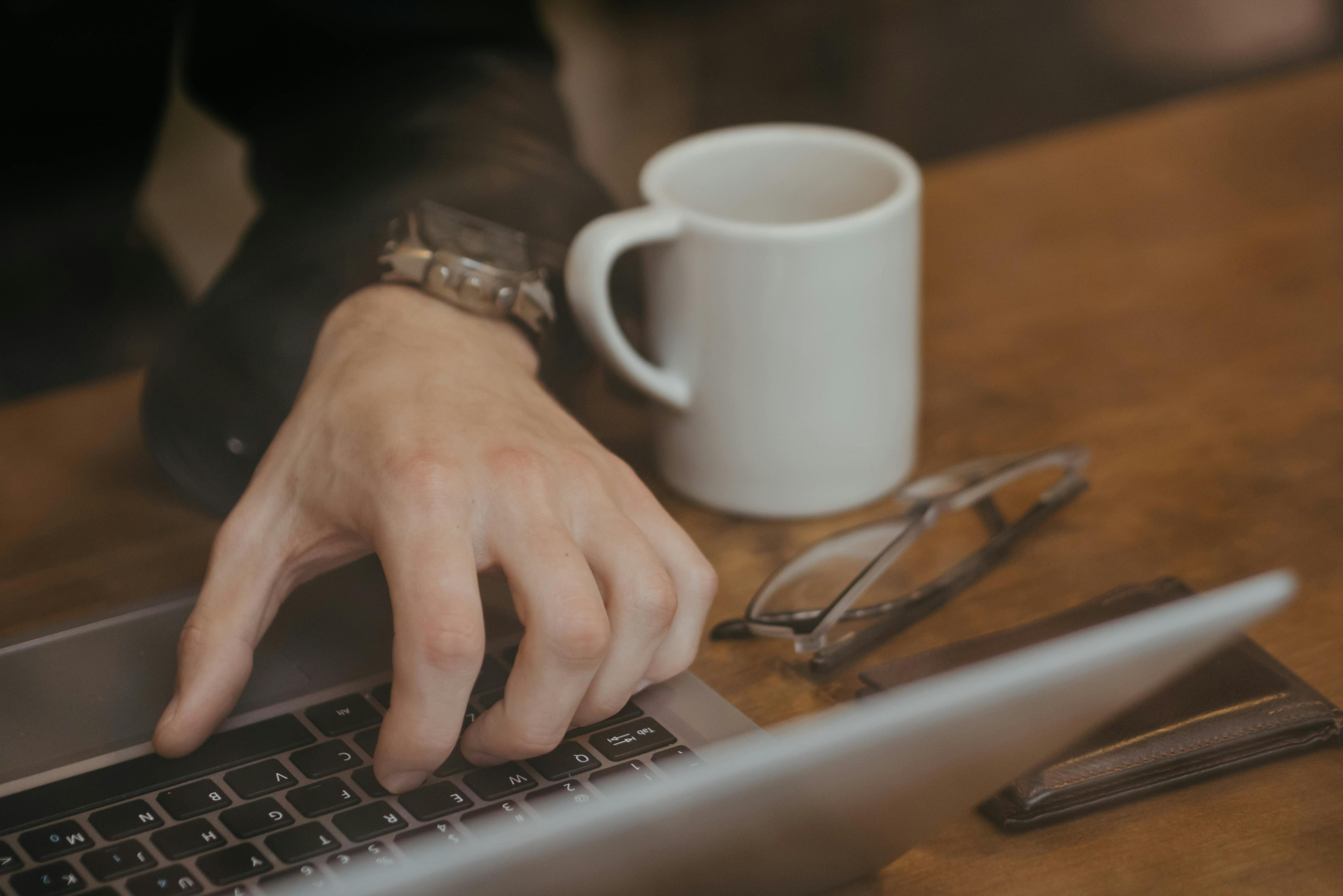 Close-up of a person working on a laptop with a coffee mug and glasses on the table.