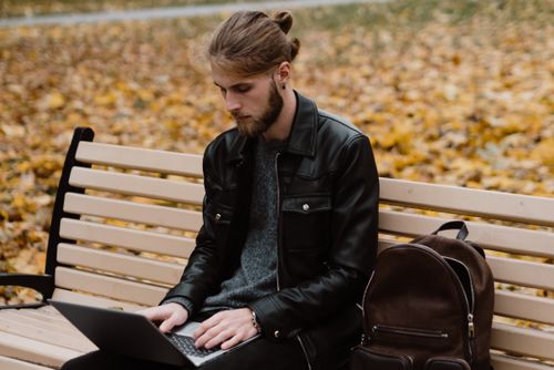 Free A man with manbun typing on a laptop while sitting on a park bench, surrounded by autumn leaves. Stock Photo
