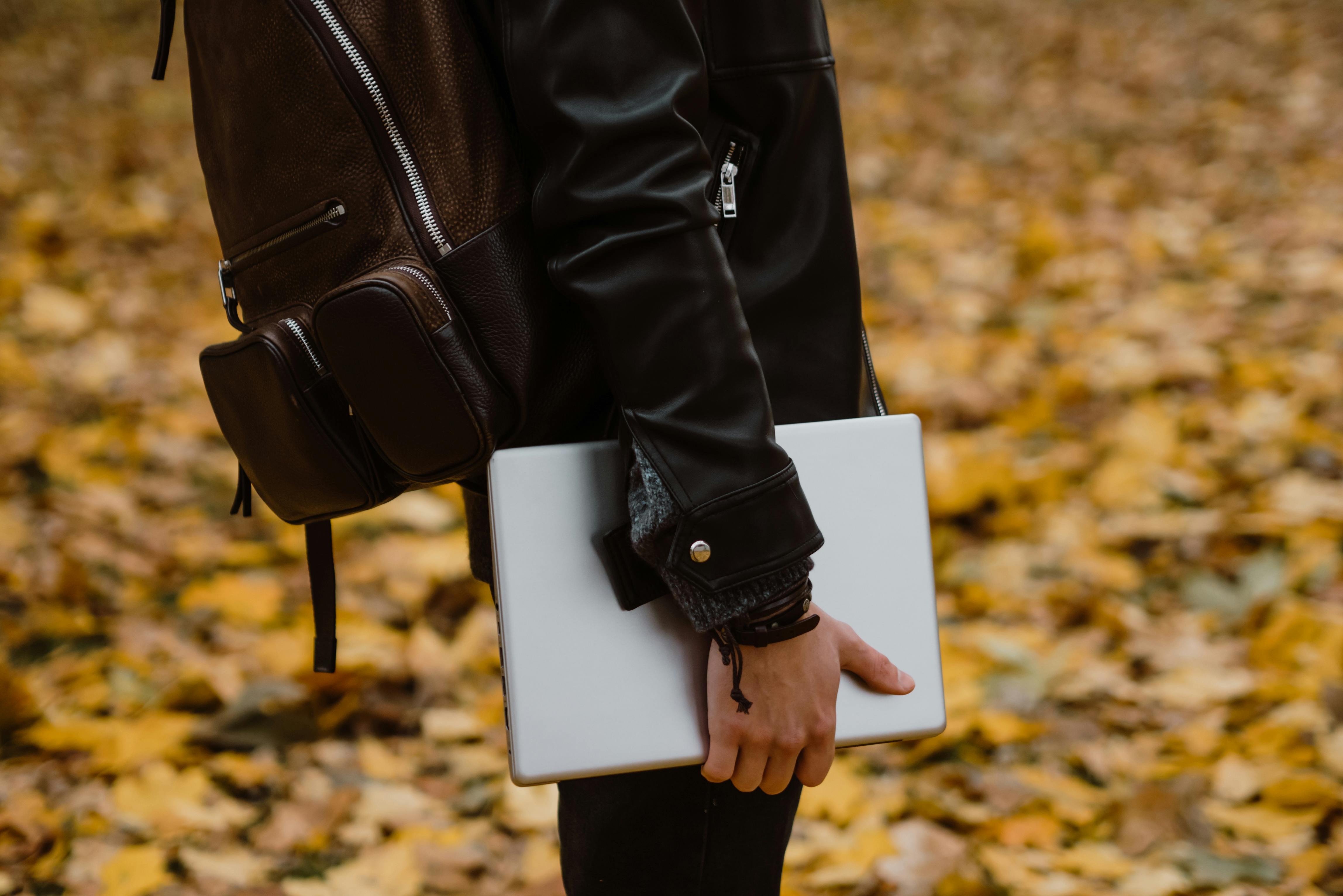 Person with backpack holding laptop amidst fallen autumn leaves, outdoors.