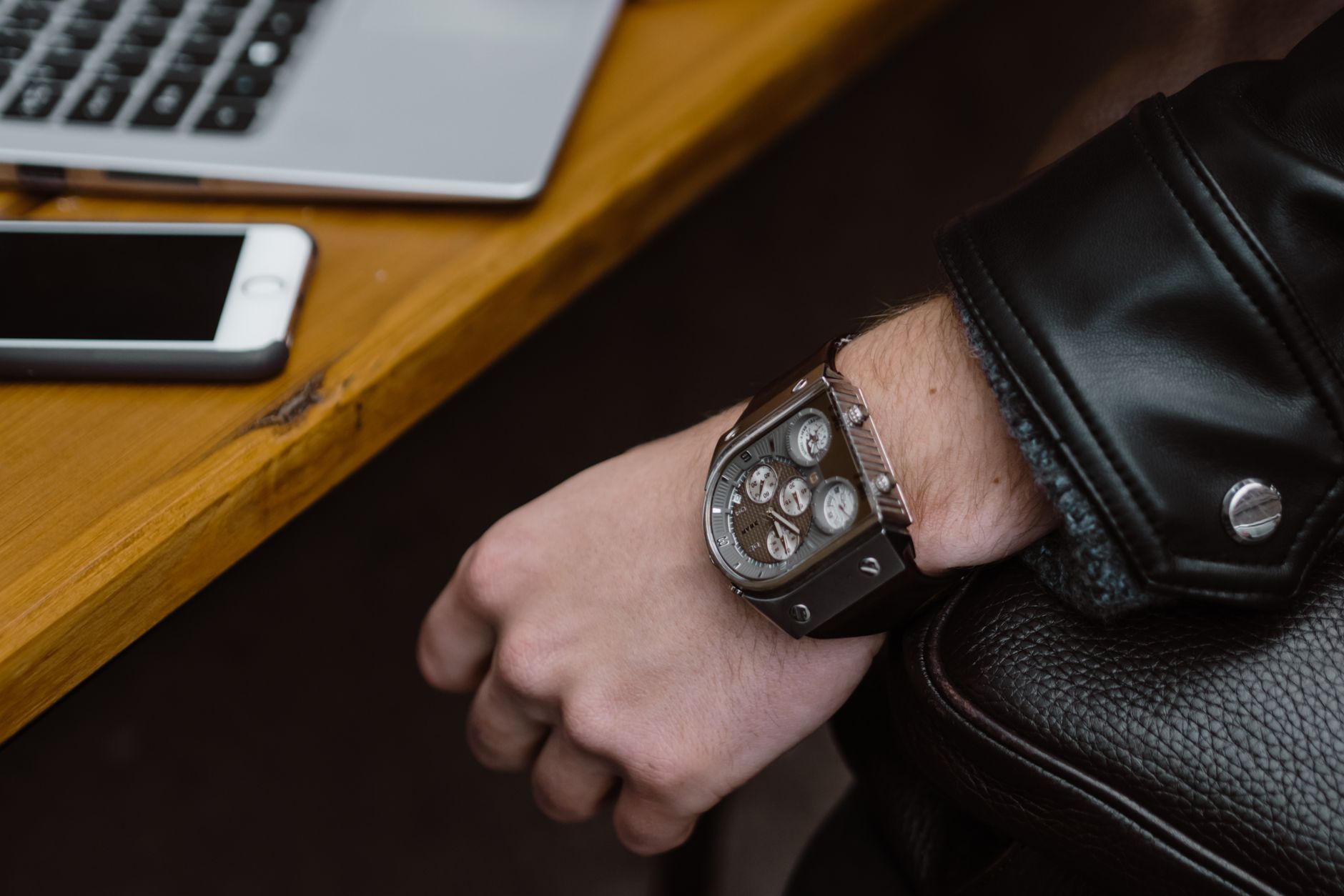 Close-up of a stylish metal wristwatch on a person's wrist with a leather jacket, next to a smartphone and laptop.