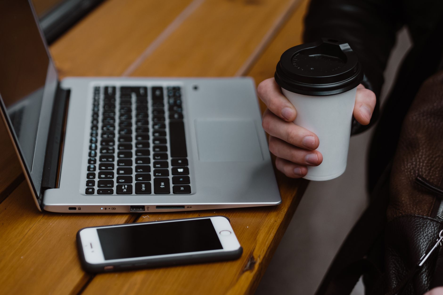 Hand holding a smartphone with a banking app, next to a cup of coffee and a laptop on a warm desk.