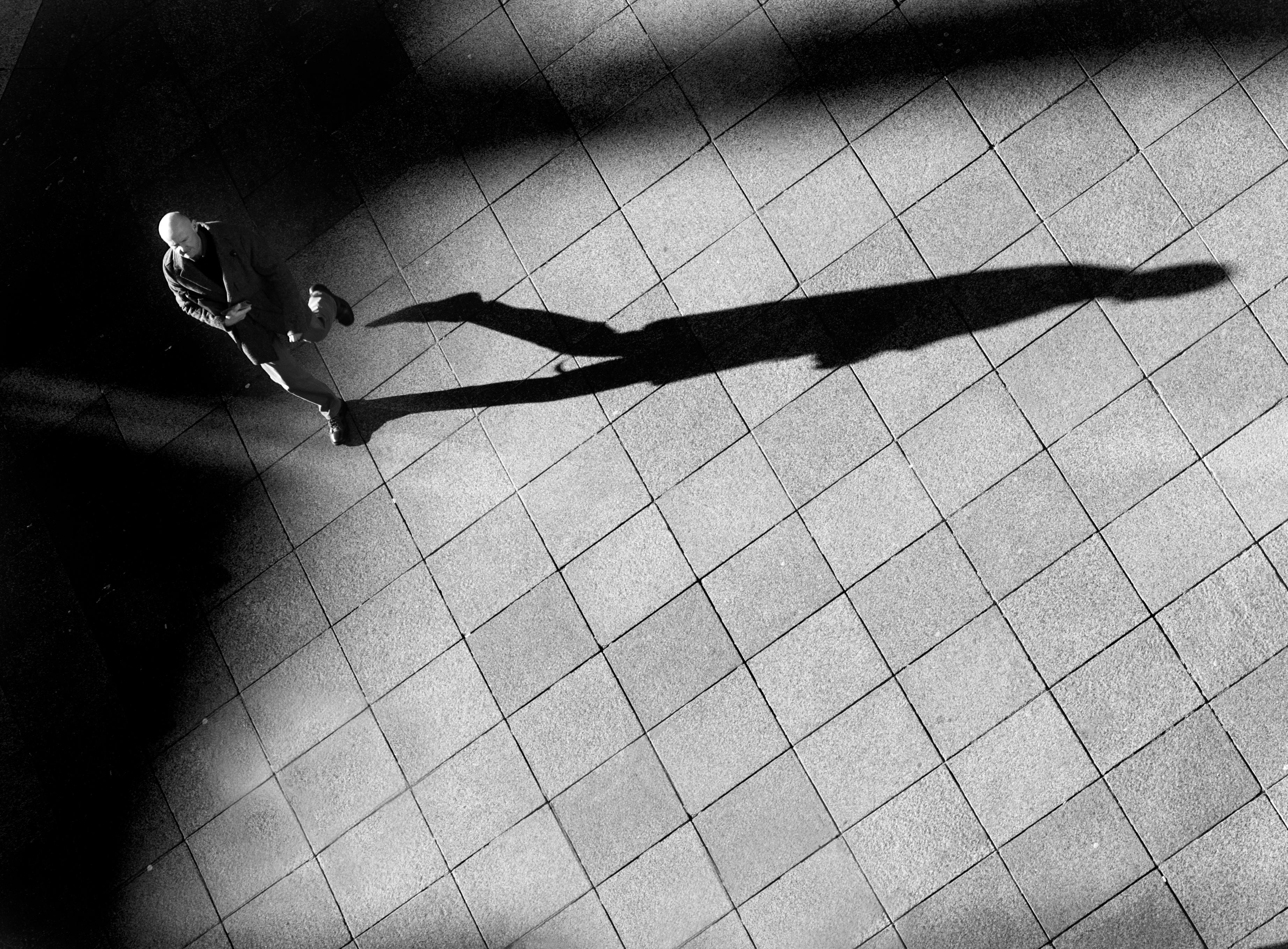 Black and white photo of a man walking on a tiled pavement casting a long shadow.