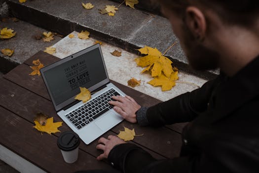 A man using a laptop surrounded by autumn leaves on a wooden bench.