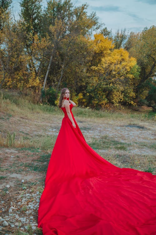 Side view of romantic female in long red dress touching hair while standing in park and looking over shoulder