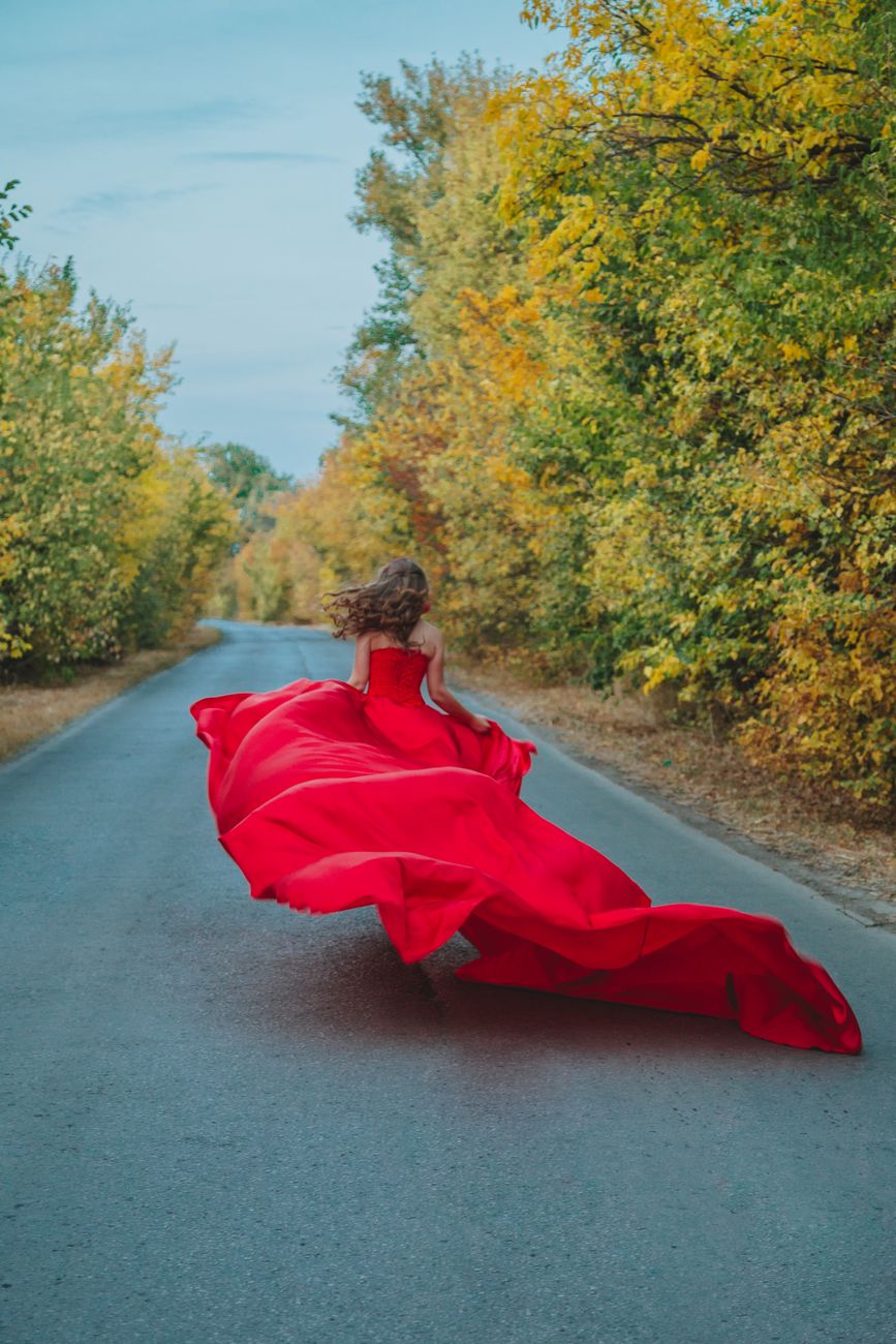 Back view of unrecognizable female in flowing long red dress running on asphalt road between autumn trees in countryside