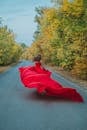 Back view of unrecognizable female in flowing long red dress running on asphalt road between autumn trees in countryside
