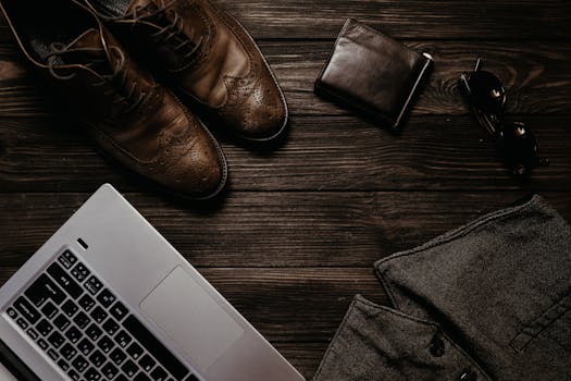 Modern workspace featuring laptop, leather shoes, wallet, and sunglasses on a wooden desk.