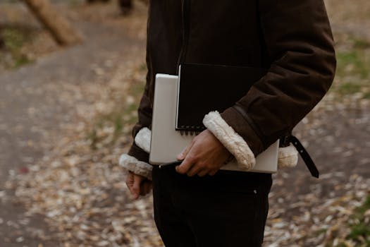 A man walks through an autumn park holding a laptop and notebook, embodying freelance lifestyle.