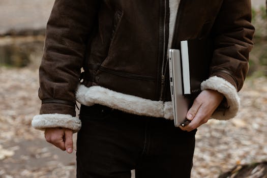 A man in a brown jacket holds a laptop and notebook in a park during fall.
