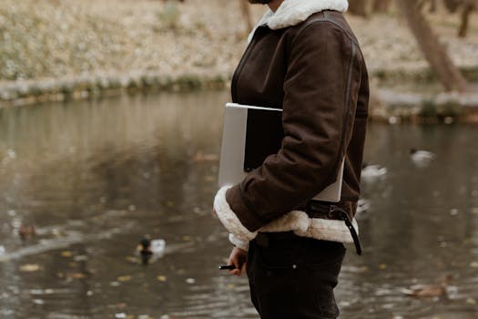 A man stands by a tranquil lake in a park, holding a laptop, surrounded by autumn leaves.