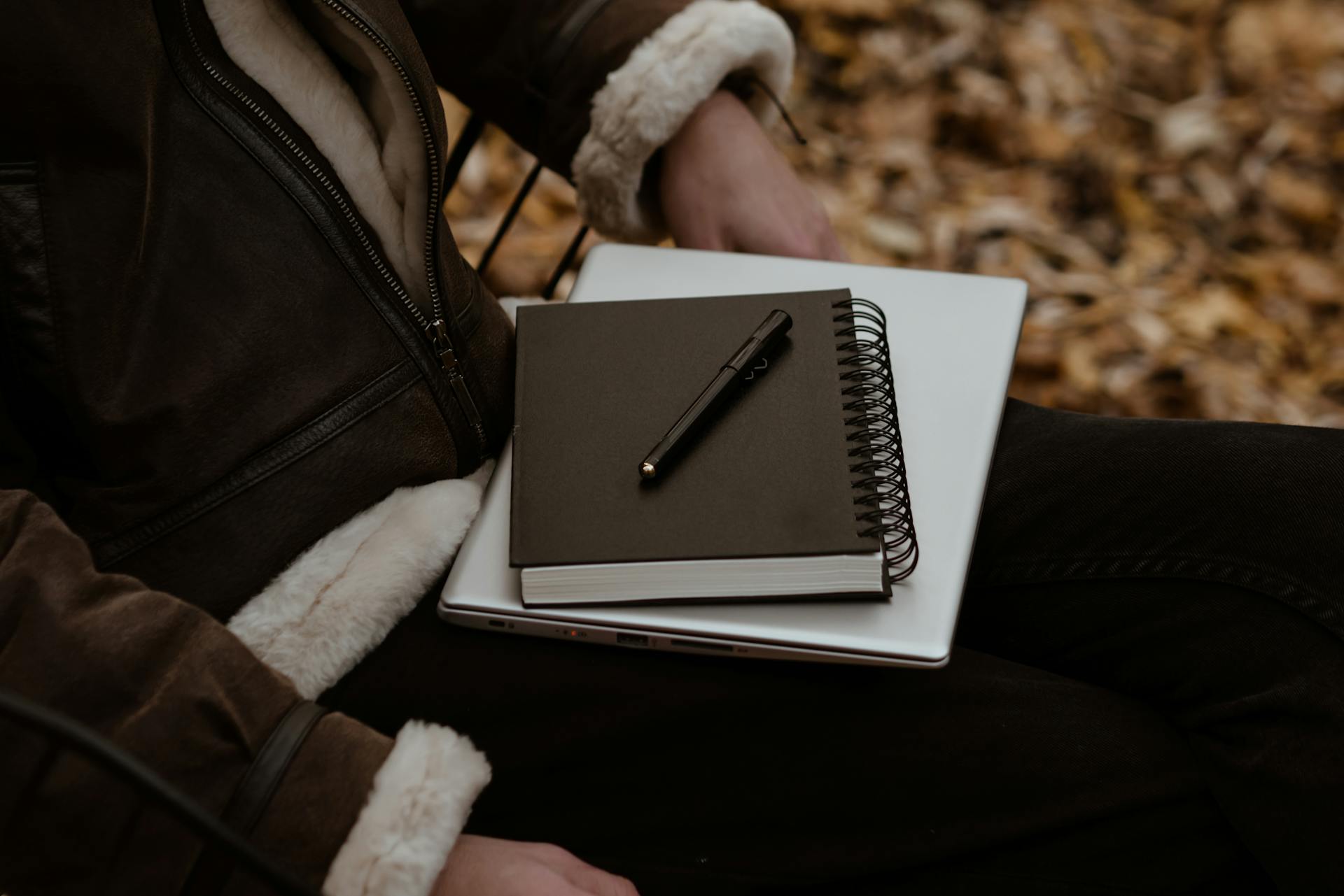 A person sitting outdoors with a notebook and laptop, surrounded by autumn leaves, representing a cozy work setup.