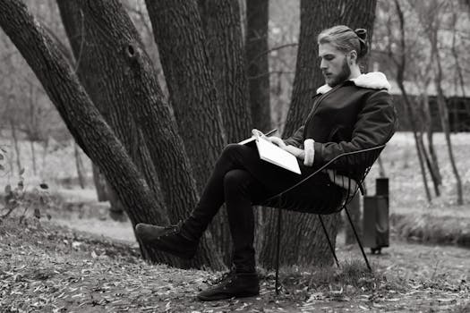 A man in a leather jacket sits on a bench in a park during fall, writing in a notebook.