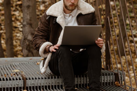 Stylish man working on laptop in an autumn park, embracing freelance lifestyle amidst natural scenery.