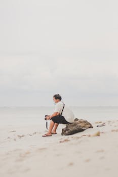 A solitary man with a camera and face mask sits on a sandy beach facing the sea.