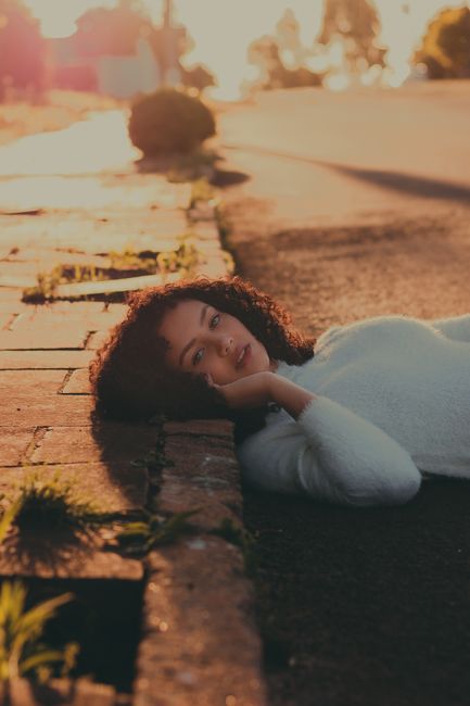 Caucasian woman with curly hair lying on urban sidewalk during sunset.